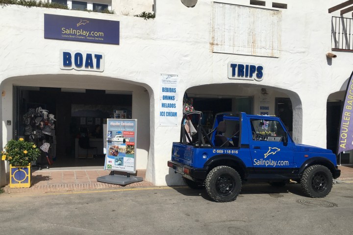 Blue jeep parked outside a shop advertising boat trips on a sunny day.