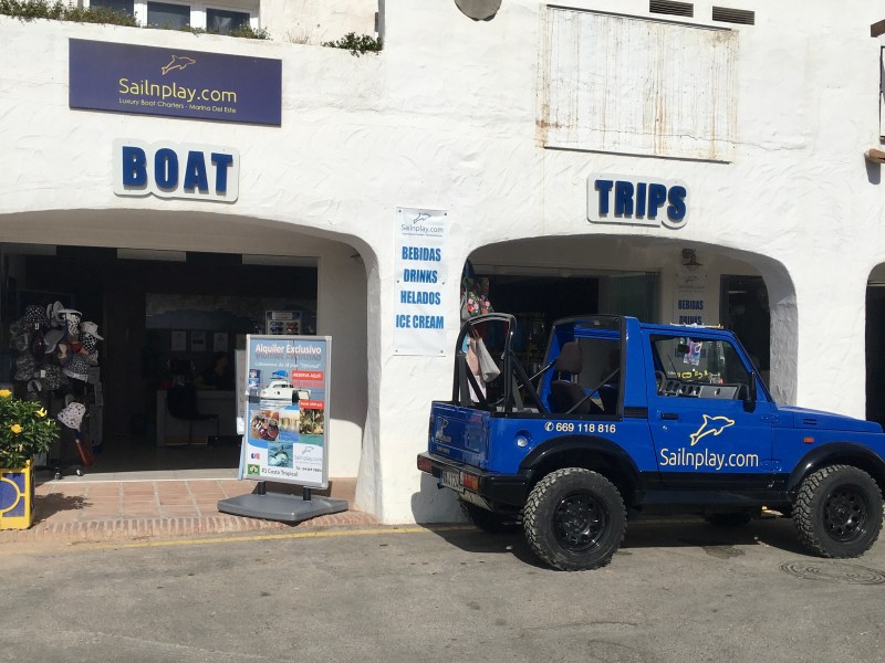 Blue jeep parked outside a shop advertising boat trips on a sunny day.