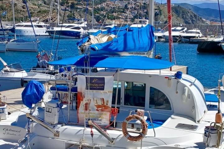 Docked sailboat with blue covers and small inflatable boat in a sunny harbor, hills and city in background.