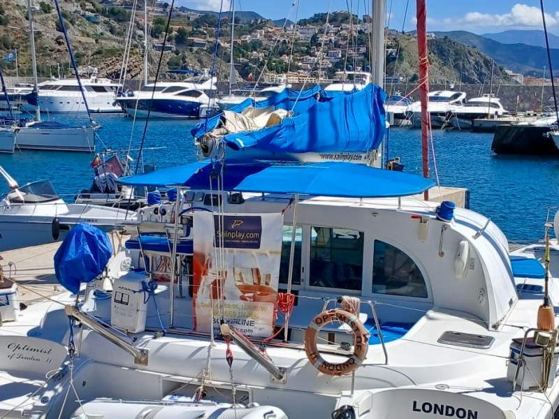 Docked sailboat with blue covers and small inflatable boat in a sunny harbor, hills and city in background.