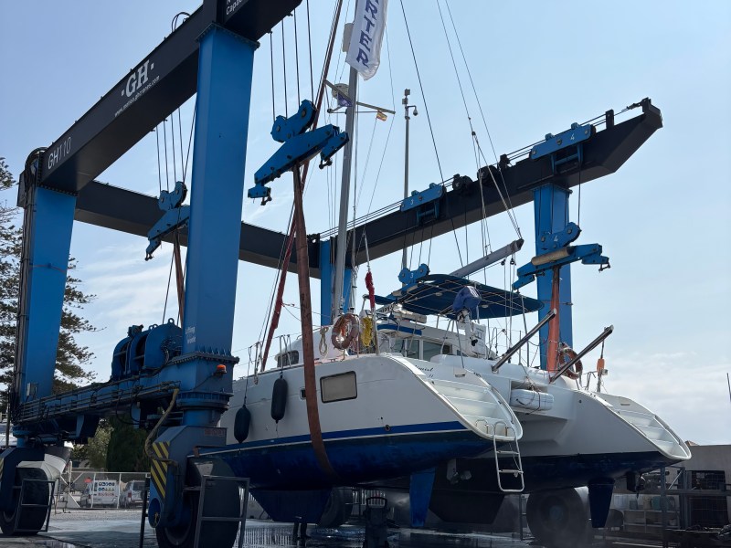 Large catamaran lifted by blue crane in boatyard under clear sky.