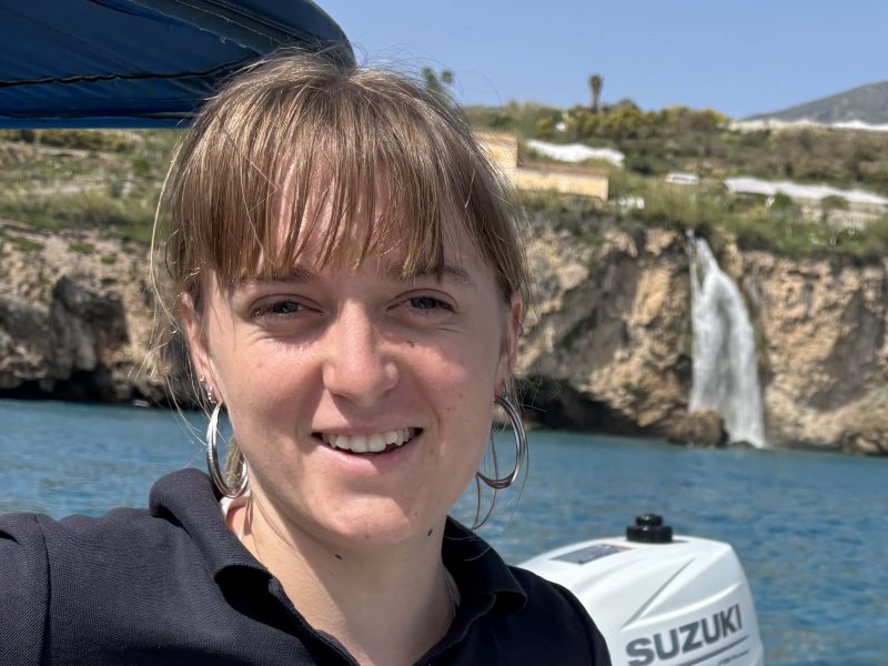 Person smiling on a boat with a waterfall and rocky coastline in the background.