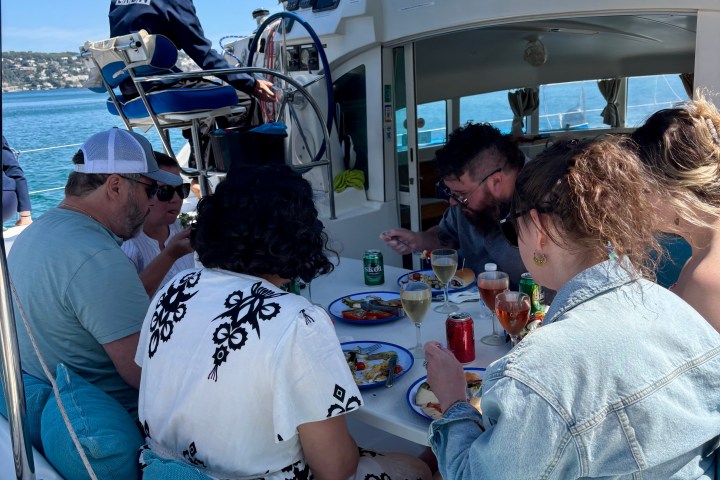 People seated on a boat, enjoying a meal with drinks, under a blue canopy, with the captain steering.