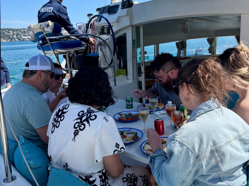 People seated on a boat, enjoying a meal with drinks, under a blue canopy, with the captain steering.
