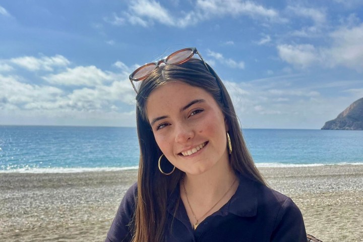 Woman smiling by the beach, wearing sunglasses and a navy shirt with 'Sailnplay.com' logo.