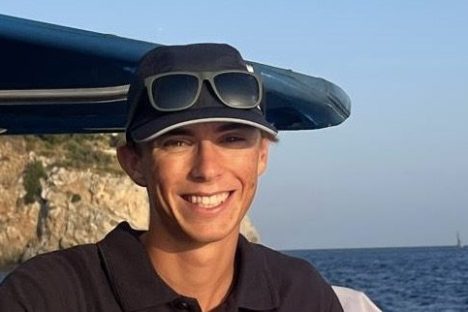 Person on a boat wearing a cap and sunglasses, with ocean and cliffs in the background.