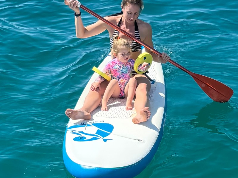 Woman and child on paddleboard in blue ocean, child holding toy, woman paddling.