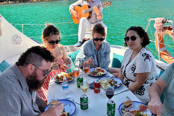 People dining on a boat deck with a guitarist playing; green water and forested shore in the background.