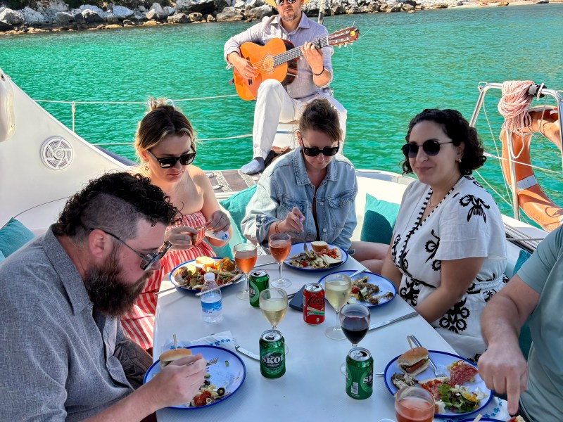 People dining on a boat deck with a guitarist playing; green water and forested shore in the background.