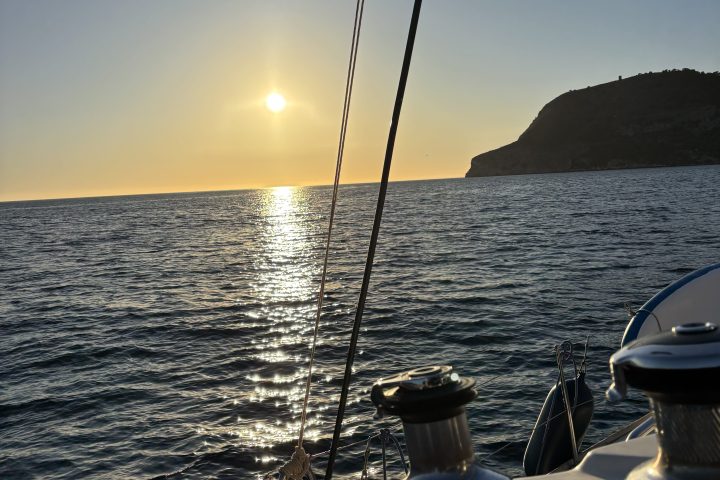 Sailing boat at sunset with calm sea and distant hillside.