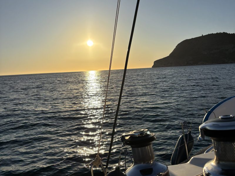 Sailing boat at sunset with calm sea and distant hillside.