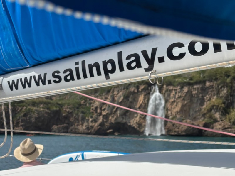 View from sailboat with sign 'sailnplay.com', cliff and waterfall in background.