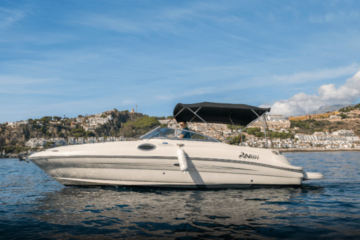 White boat with canopy on water, hillside buildings in background under blue sky.