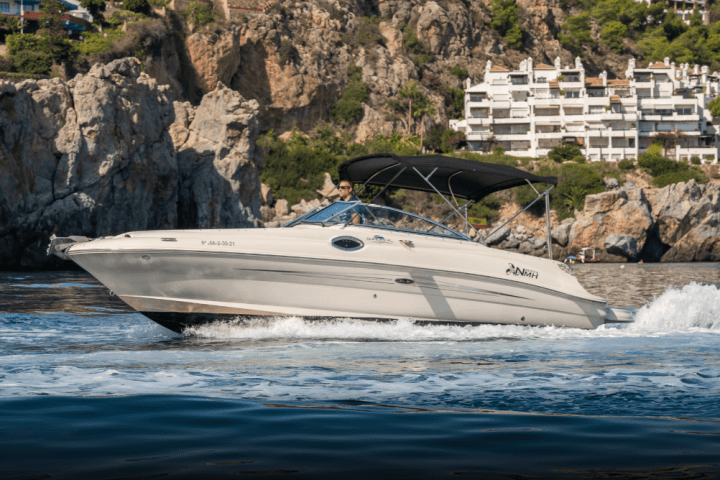 Speedboat cruising near rocky coastline and white buildings under clear sky.