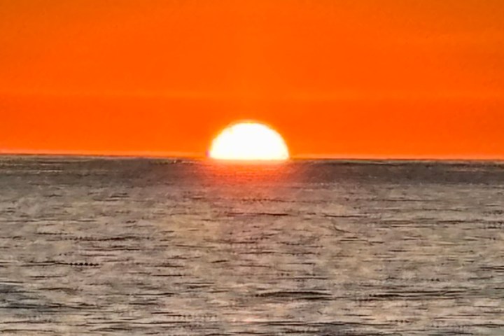 Sunset over ocean with orange sky and sun half-visible above horizon.